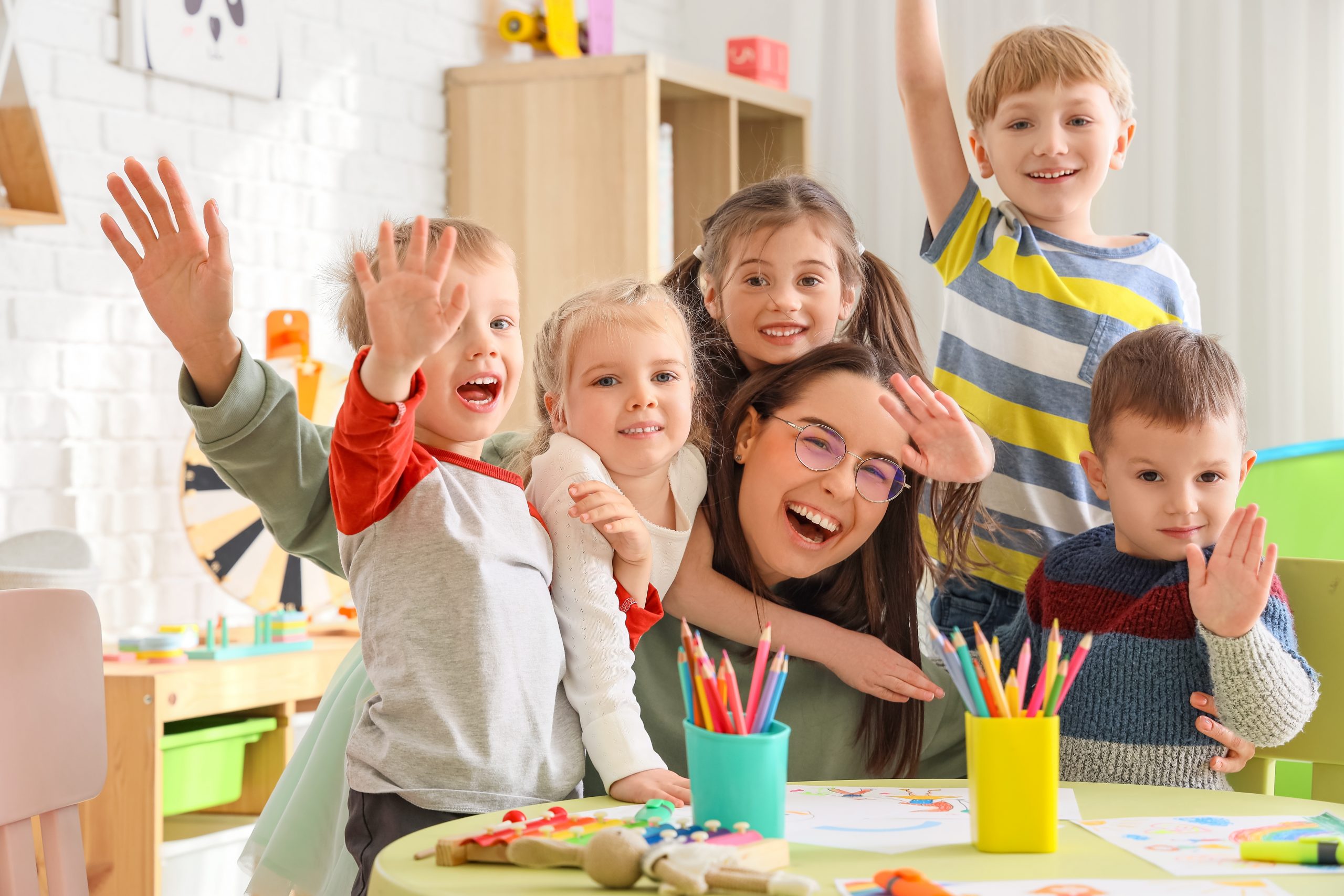 Preschool kids with their teacher waving at a table filled with arts and crafts in a classroom.